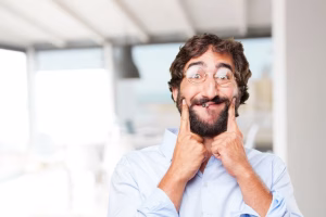 A man with a beard and glasses is putting his finger on his nose.